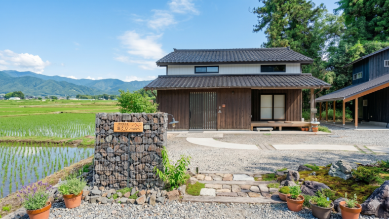 A panoramic 16:9 view of "Asahi Sado," a modern Japanese guest house set against a peaceful rural landscape. The image features a traditional dark-wood house with a tiled roof, a lush green rice paddy field on the left, and a backdrop of rolling mountains under a clear blue sky. In the foreground, a wooden sign reading "Asahi Sado" is mounted on a gabion stone wall, with no power lines obstructing the view.