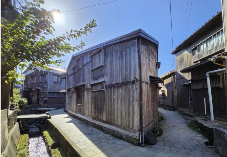 The weathered wood, the narrow stone-paved lane, and the water channel read as authentic and lived-in rather than a polished stock photo — exactly the tone your article needs. No people, no cars, no signage, so it's clean for professional use. The sun flare in the corner adds warmth rather than distraction.