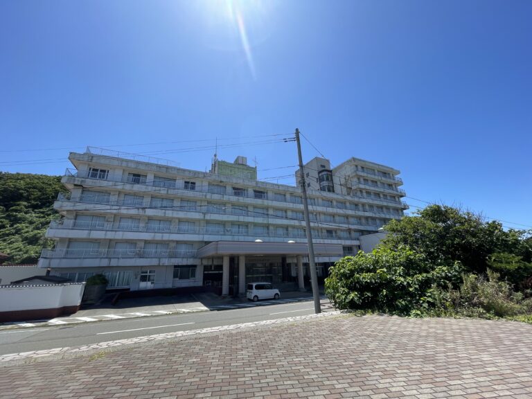 A multi-story hotel building with white balconies, under a clear blue sky, surrounded by trees.